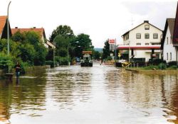 In der Mitte des Bildes die von Hochwasser überflutete Straße auf der ein Traktor von hinten zu sehen ist. Links und rechts davon Bebauung.